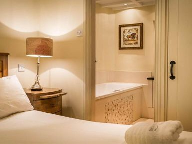 A bedroom with a wooden nightstand and lamp next to a view into a bathroom with a bathtub and lion picture at Ivy Cottage in Ambleside