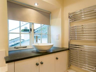 A bathroom sink with a round basin on a dark countertop under a window with a shade and a chrome towel heater on the wall at Ivy Cottage in Ambleside