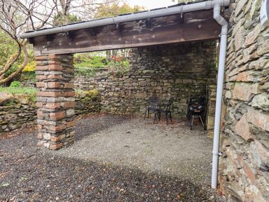 An outdoor covered seating area with stone walls, two black metal chairs, a charcoal grill, and gravel ground at The Old Chapel in Crook