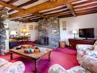 A living room with a stone fireplace wooden beams floral armchairs a wooden coffee table and a dining table at Bank Barn at Rusland Grizedale
