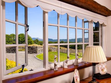 A window with white frame and scalloped valance overlooking a garden and hills with ceramic figurines and a table lamp on the windowsill at Bank Barn at Rusland Grizedale