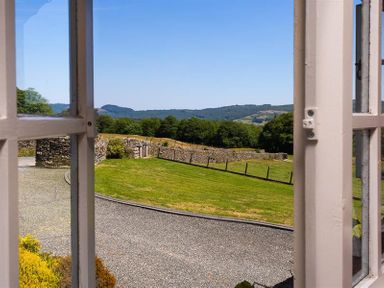 A view of a gravel driveway and a fenced grassy area with hills and trees beyond seen through an open window at Bank Barn at Rusland in Grizedale