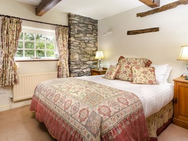 A bedroom with a bed covered in patterned bedding two bedside tables with lamps a window with floral curtains and a stone wall Bank Barn at Rusland Grizedale