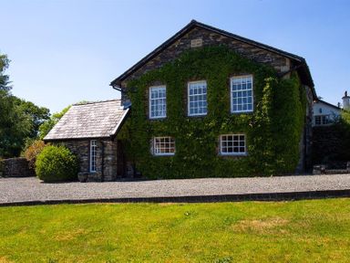 A stone house with ivy on the walls and multiple windows with a gravel driveway and grass lawn at Bank Barn at Rusland in Grizedale