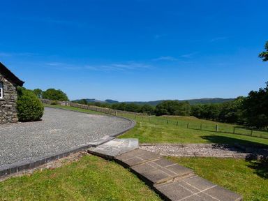A gravel driveway next to a stone building with a curved stone pathway and green fields in the background at Bank Barn at Rusland Grizedale