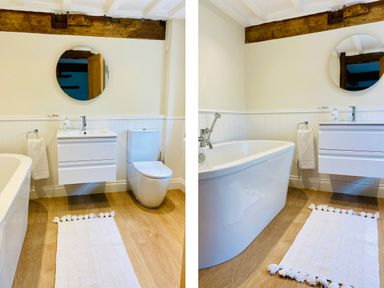 A bathroom with a white bathtub a white sink with cabinet a round mirror a toilet and a white rug on wooden floor at Bank Barn at Rusland in Grizedale