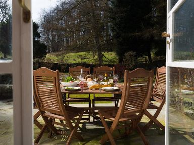 A wooden outdoor dining table set with plates wine and bread on a patio in a garden