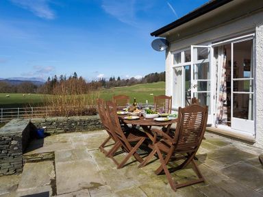 An outdoor patio with a wooden dining table set for a meal next to a white house with open glass doors at Juniper Cottage in Windermere
