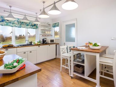 A kitchen with white cabinets wooden countertops a small dining table with chairs and large windows showing a rural view at Juniper Cottage in Windermere