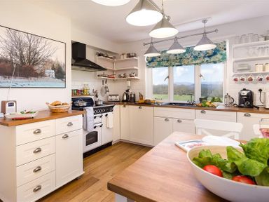 A kitchen with white cabinets a stove a window with a floral blind and a bowl of salad on the table at Juniper Cottage in Windermere