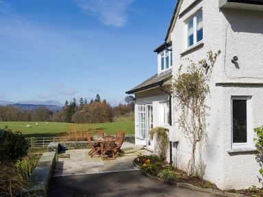 A white house with a patio table and chairs outside overlooking a green field with sheep at Juniper Cottage in Windermere