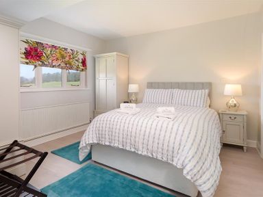 A bedroom with a bed two bedside tables with lamps a wardrobe window with floral blinds and blue rugs at Juniper Cottage in Windermere