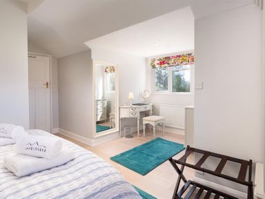 A bedroom with a bed, a full-length mirror, a dressing table with a stool, and a window with a floral curtain at Juniper Cottage Windermere