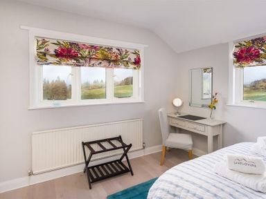 A bedroom with two windows with floral curtains a desk with a chair and mirror and a luggage rack at Juniper Cottage in Windermere