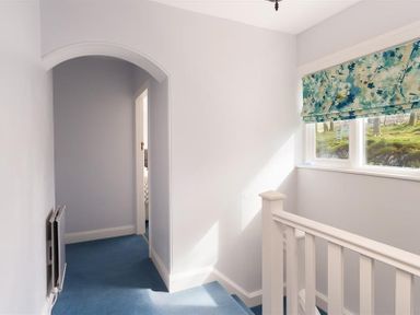 A hallway with blue carpet a radiator an arched doorway and a window with floral curtains at Juniper Cottage in Windermere