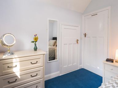 A bedroom corner with a dresser with a round mirror and vase with flowers a full length mirror two closed doors and a small bedside table with a lamp and alarm clock at Juniper Cottage in Windermere
