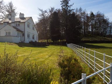 An outdoor view of a white house with a fenced grassy yard and trees in the background at Juniper Cottage in Windermere