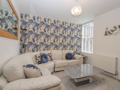 A living room with a white sectional sofa blue patterned and round pillows a glass coffee table patterned wallpaper and a window with blinds at Rossett Holme in Ambleside