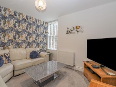 A living room with a beige leather sofa blue patterned cushions floral wallpaper a grey coffee table and a wooden TV stand with a television