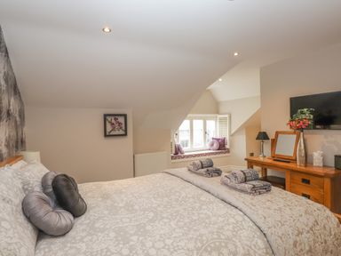 A bedroom with a bed, a window seat with cushions, a wooden dresser with flowers, a mirror, and a television at Rossett Holme in Ambleside