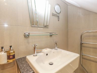 A bathroom sink with two taps a soap dish a small wall mirror a glass shelf and bottles on a countertop at Rossett Holme in Ambleside