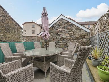 An outdoor patio with a glass table surrounded by eight wicker chairs and a closed umbrella at Rossett Holme in Ambleside