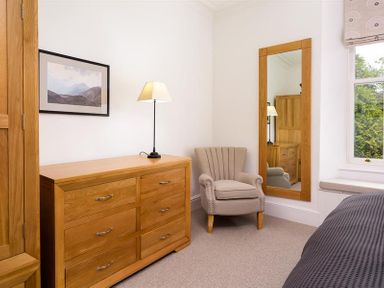 A bedroom with a wooden dresser lamp armchair wall mirror and window at Ben House in Grasmere