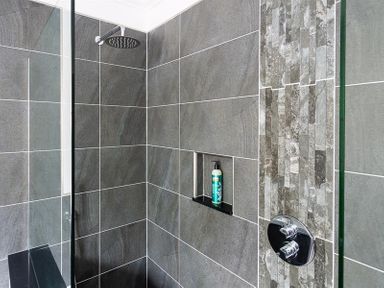 A shower area with grey tiles a built in shelf holding a bottle and a glass shower screen at Ben House in Grasmere