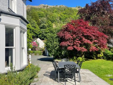 An outdoor patio with a table and six chairs next to a white house and garden with trees at Ben House in Grasmere