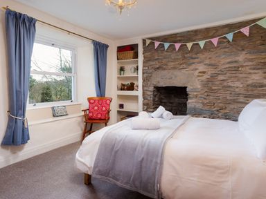 A bedroom with a bed rolled towels a stone fireplace a wooden chair with a cushion blue curtains and a window at Birchmill Cottage Crook
