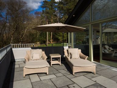 Two patio loungers with cushions under a large umbrella on a stone terrace outside a building with glass doors at Tall Trees in Bowness