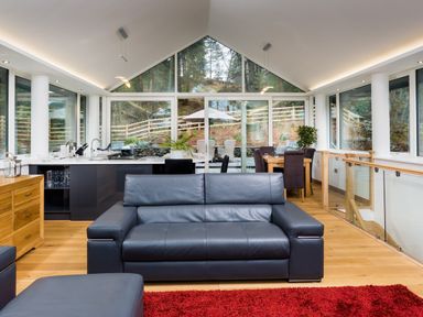 A living room with a black leather sofa and large windows overlooking a wooden deck at Tall Trees in Bowness