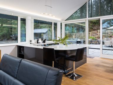 A modern kitchen with black chairs and white island countertop in a room with large windows and wooden floor at Tall Trees in Bowness