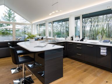 A modern kitchen with black cabinets white countertops bar stools and large windows overlooking trees at Tall Trees in Bowness