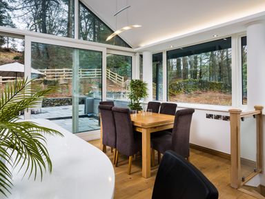 A dining area with a wooden table and six chairs next to large windows showing an outdoor patio and wooded area at Tall Trees in Bowness