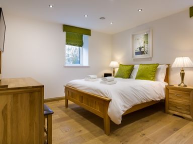 A bedroom with a wooden bed with white bedding and green pillows two bedside tables with lamps and a wooden dresser at Tall Trees in Bowness