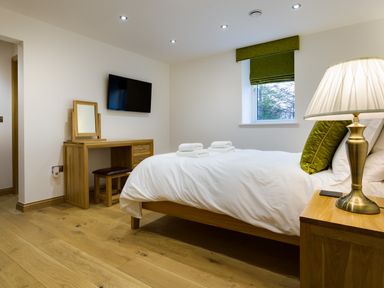 A bedroom with a wooden bed with white bedding and green cushions next to a wooden nightstand with a lamp and a wooden dressing table with a mirror and stool and a wall-mounted television at Tall Trees in Bowness
