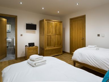 A bedroom with two beds and white linens a wooden wardrobe and bedside table a small wall-mounted television and an open door leading to a bathroom at Tall Trees in Bowness