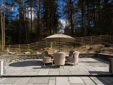 An outdoor patio with a round table umbrella and six wicker chairs surrounded by a stone wall and wooden fence at Tall Trees in Bowness