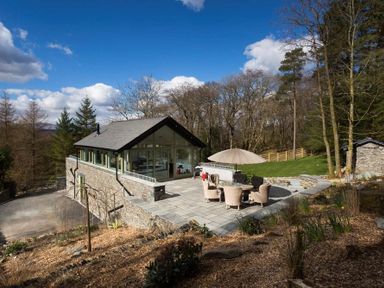 An outdoor patio area with wicker chairs around a round table and an umbrella next to a stone building surrounded by trees at Tall Trees in Bowness