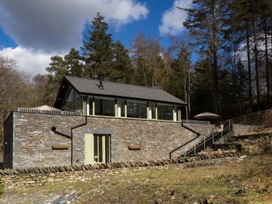 A stone house with large windows next to a forest and stairs at Tall Trees in Bowness