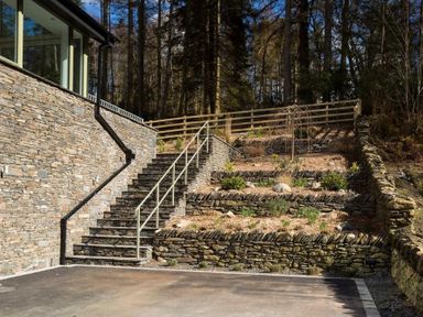 Stone stairs and terraced garden beds with plants beside a stone wall and forest at Tall Trees in Bowness