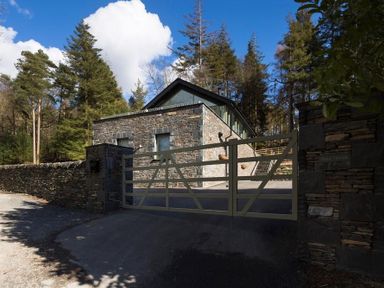 A stone house with a gated driveway surrounded by trees at Tall Trees in Bowness