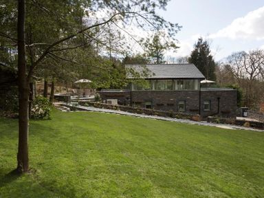 A house with large windows and a patio with chairs and umbrellas surrounded by grass and trees at Tall Trees in Bowness