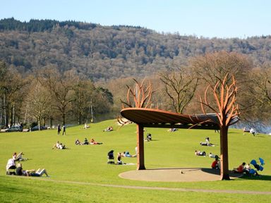 A park with people sitting on grass near a shelter with tree-like metal structures in the background at Tall Trees in Bowness