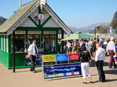 A ticket booth with people standing near a sign for lake cruises at Pier 2 with hills in the background at Tall Trees in Bowness