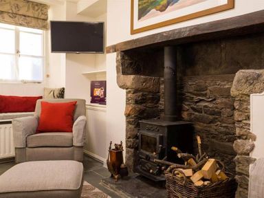 A sitting room with a stone fireplace and wood stove a basket of firewood an armchair with a red cushion and a wall-mounted television at Lettera in Ambleside