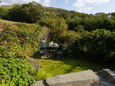 A small garden area with green plastic chairs around a table set with bowls and fruits surrounded by dense greenery and hills in the background at Lettera Ambleside