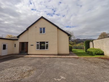 A cream house with a gravel driveway and a small garden with trees and mountains in the background at Laal Field House in Keswick