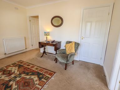 A room with a patterned rug chair with a yellow cushion small wooden table with a lamp and a round mirror on the wall at Laal Field House in Keswick
